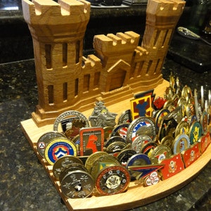 Puede incluir: Expositor de madera con forma de fuerte militar, con ranuras para guardar una colecci&oacute;n de monedas de desaf&iacute;o militares. Las monedas son principalmente de oro y plata con varios dise&ntilde;os y logotipos.