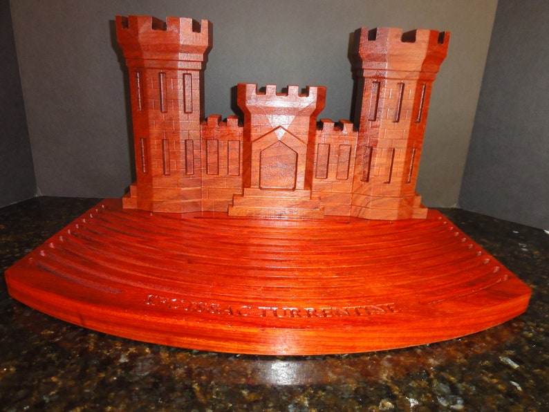 Puede incluir: Un modelo de madera de un castillo con un acabado rojo. El castillo tiene dos torres y una puerta central. El modelo est&aacute; sentado sobre una base de madera con un borde curvo. La base tiene una serie de c&iacute;rculos conc&eacute;ntricos tallados en ella. La base est&aacute; inscrita con las palabras "Corps of Engineers".