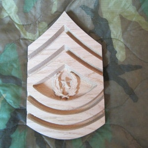 Puede incluir: Un recorte de madera de una insignia de rango militar, con un diseño de chevron con tres rayas y un emblema central. La insignia está hecha de madera de color claro y tiene un acabado liso y pulido.