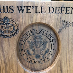 May include: A wooden plaque with the words "THIS WE'LL DEFEND" engraved on it. The plaque also features a circular design with the words "UNITED STATES ARMY" and a stylised eagle. There is a smaller circular design with the words "THIS WE'LL DEFEND" and a stylised military insignia. The plaque also features a pair of crossed pistols.