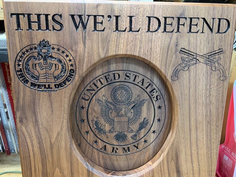May include: A wooden plaque with the words "THIS WE'LL DEFEND" engraved on it. The plaque also features a circular design with the words "UNITED STATES ARMY" and a stylised eagle. There is a smaller circular design with the words "THIS WE'LL DEFEND" and a stylised military insignia. The plaque also features a pair of crossed pistols.