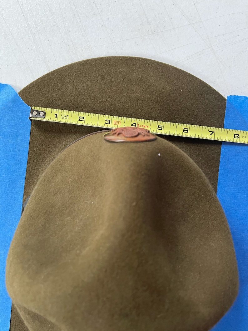 May include: A green felt hat with a brown leather band and a metal badge that says "Lufkin" on it. The hat is being measured with a tape measure that reads 7 inches.