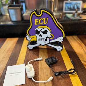 May include: A wooden pirate-themed sign featuring the ECU logo, with a skull and crossbones design. Includes a white dimmer switch, a black power adapter, and a user instruction manual for LED strip lights.