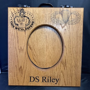 May include: A wooden box with a cutout for a picture or medal. The box has a black handle and two latches. The box is engraved with the words "This We'll Defend" and a military insignia. The box also has a paratrooper insignia and the name "DS Riley" engraved on it.