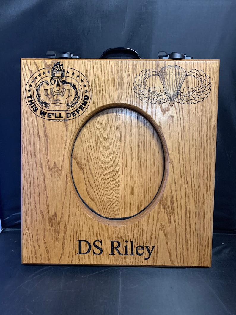 May include: A wooden box with a cutout for a picture or medal. The box has a black handle and two latches. The box is engraved with the words "This We'll Defend" and a military insignia. The box also has a paratrooper insignia and the name "DS Riley" engraved on it.