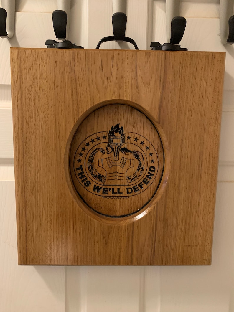 May include: A wooden box with a hinged lid and a carved design of a military insignia. The insignia features a torch, a wreath, and the text "This We'll Defend".
