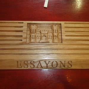 May include: A rectangular wooden desk organizer with a carved architectural design and the word "ESSAYONS" engraved on the front. The organizer has multiple slots and a natural wood finish.