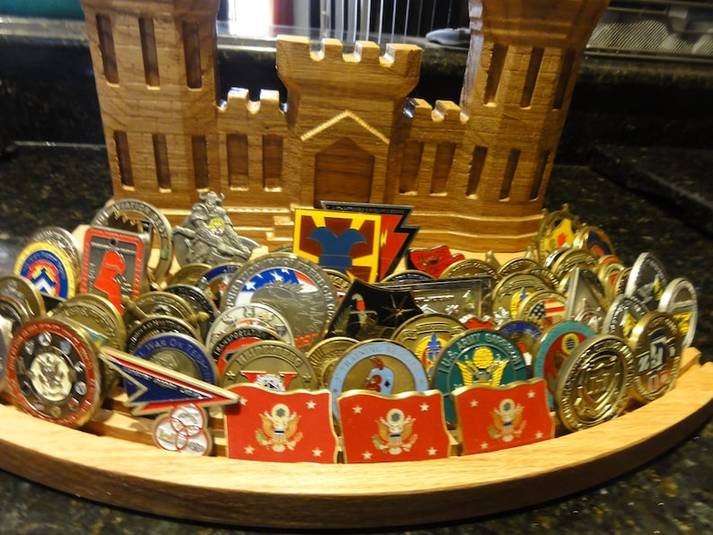 Puede incluir: Un expositor de madera con forma de fortaleza con una colecci&oacute;n de monedas militares de desaf&iacute;o. Las monedas est&aacute;n dispuestas en semic&iacute;rculo y presentan diversos dise&ntilde;os, incluyendo banderas, emblemas e insignias militares.