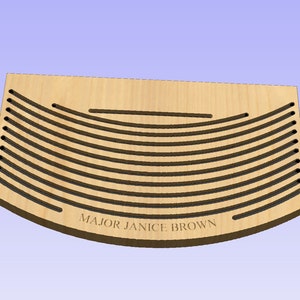 Puede incluir: Una placa de madera con forma curva y una serie de l&iacute;neas paralelas cortadas en la superficie. La placa tiene el texto "MAJOR JANICE BROWN" grabado en el borde inferior.