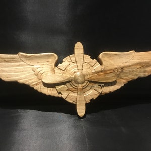 以下が含まれることがあります： 中央にプロペラが付いた木製の彫刻のパイロットの翼。翼は木目模様で詳細に作られており、プロペラはリアルなデザインで彫刻されています。