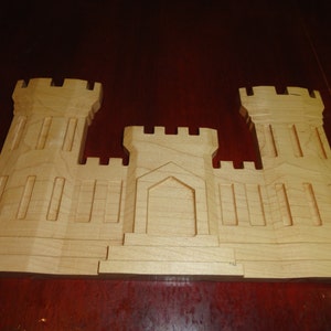 May include: A wooden model of a castle with two towers and a central section. The model is made of light brown wood and has a simple, rustic design.