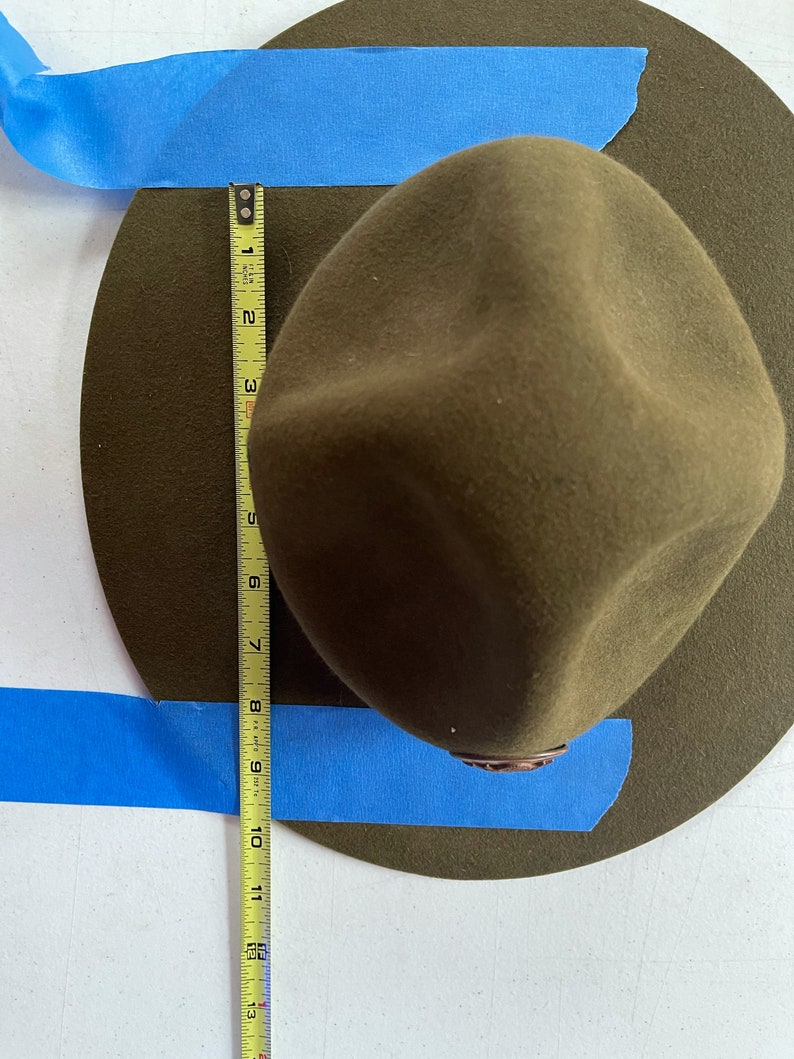 May include: A green felt hat with a brown leather band. The hat is being measured with a yellow tape measure, which shows a measurement of 10 inches. The hat is surrounded by blue painter's tape.