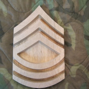 Puede incluir: Una insignia de rango militar de madera de color marrón claro con tres galones. La insignia tiene forma de galón y tiene una superficie lisa y pulida.