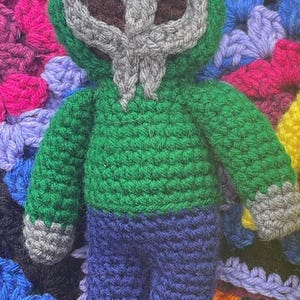 May include: A handmade crochet doll with a grey mask and green hooded top, blue trousers, and grey cuffs. The doll is set against a colourful, crocheted background. The doll's mask has brown eye holes.