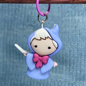 May include: A charm featuring a cartoon fairy in a light blue hooded gown with a pink bow and a wand. The charm is attached to a purple ring with a silver clasp. The fairy has white hair and black eyes.