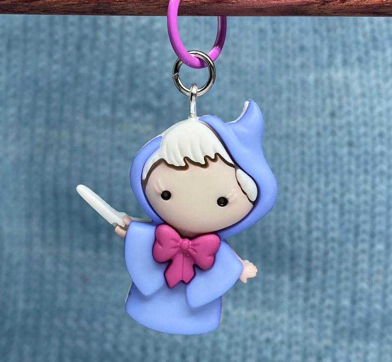 May include: A charm featuring a cartoon fairy in a light blue hooded gown with a pink bow and a wand. The charm is attached to a purple ring with a silver clasp. The fairy has white hair and black eyes.