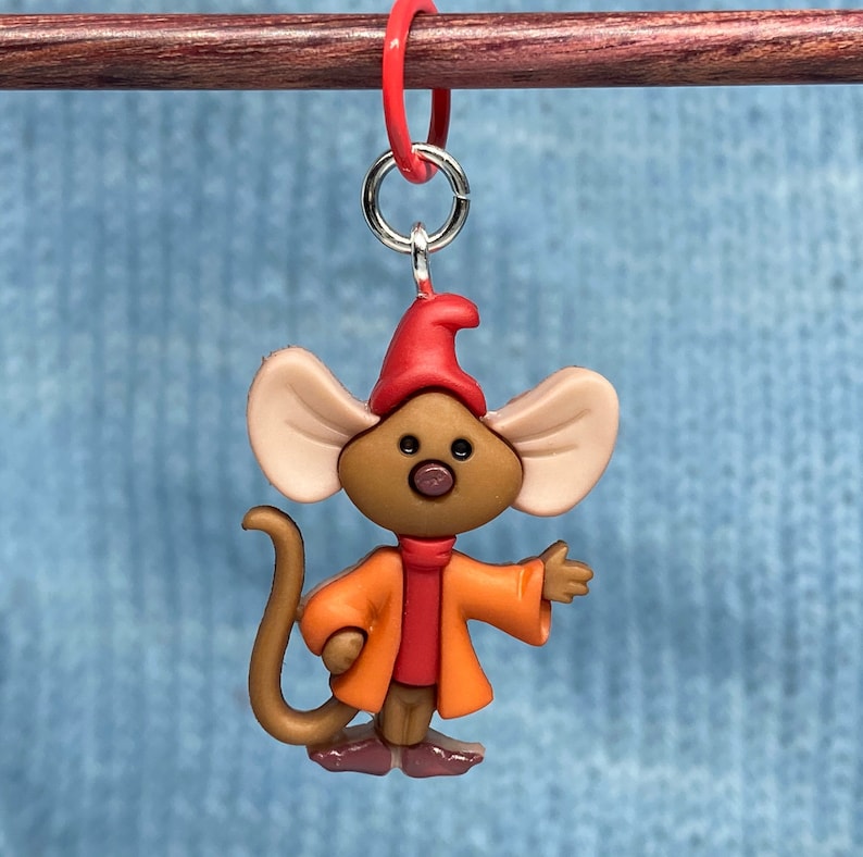 May include: A whimsical stitch marker featuring a cartoon mouse character in a red hat and orange coat. The mouse has large ears and is attached to a silver ring and a red plastic loop, perfect for knitting or crochet projects.