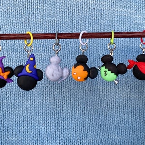 May include: A collection of six Halloween-themed stitch markers. Each marker features a black mouse head with different costume designs, including a witch hat, wizard hat, mummy, pumpkin, Frankenstein, and pirate. The markers are attached to a wooden knitting needle.