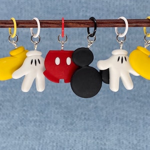 May include: Six stitch markers with cartoon character designs. The markers include yellow gloves, white gloves, a red character head, and a black character head. Each marker hangs from a silver ring.