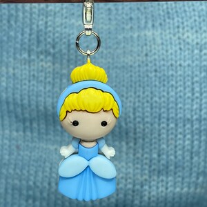 May include: A Cinderella-themed charm featuring a cartoon-style design. The charm has a light blue dress, blonde hair, and a yellow crown. It is attached to a silver-colored clasp.