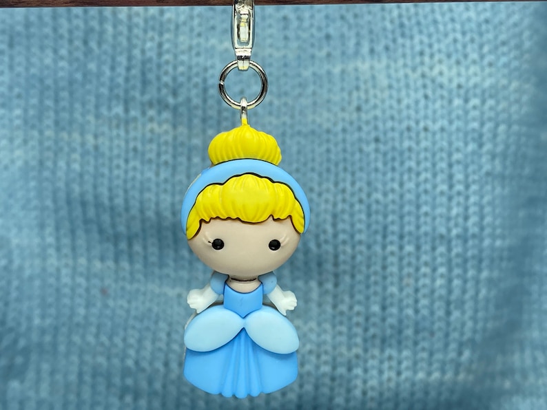 May include: A Cinderella-themed charm featuring a cartoon-style design. The charm has a light blue dress, blonde hair, and a yellow crown. It is attached to a silver-colored clasp.