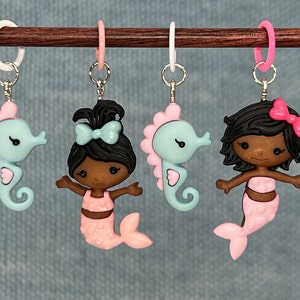 May include: Four stitch markers with mermaid and seahorse charms. The charms are in pastel colors of pink and blue, with black hair and silver-colored metal findings. The stitch markers are attached to a wooden knitting needle.