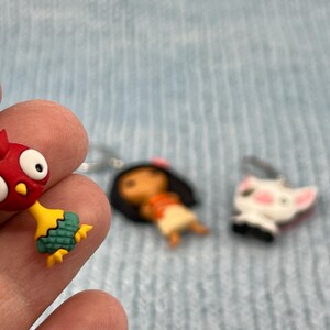 Puede incluir: Un llavero de gallo rojo y amarillo con cola verde, un llavero de ni&ntilde;a de pelo casta&ntilde;o con vestido rojo y blanco, y un llavero de perro blanco con mejillas rosas.