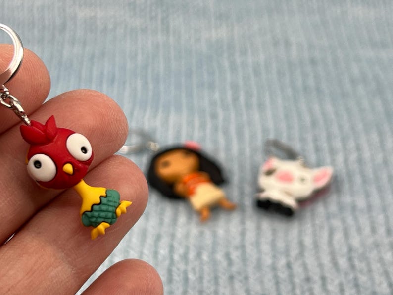 Puede incluir: Un llavero de gallo rojo y amarillo con cola verde, un llavero de ni&ntilde;a de pelo casta&ntilde;o con vestido rojo y blanco, y un llavero de perro blanco con mejillas rosas.