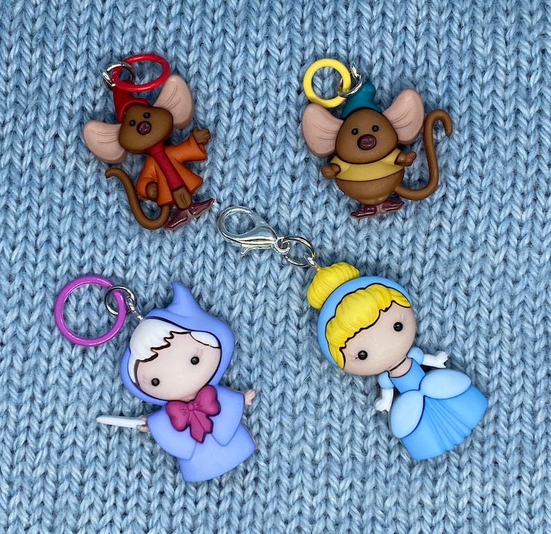 May include: Four colorful charms featuring characters from a fairy tale. Two charms are mice wearing outfits, one is a fairy with a wand, and the last is a princess in a blue dress. Each charm has a metal ring or clasp.