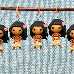 Puede incluir: Cinco marcadores de punto de ganchillo coloridos con un personaje de dibujos animados con cabello casta&ntilde;o, un corpi&ntilde;o marr&oacute;n, una falda amarilla y una flor rosa. Los marcadores de punto est&aacute;n colgados de una varilla de madera.