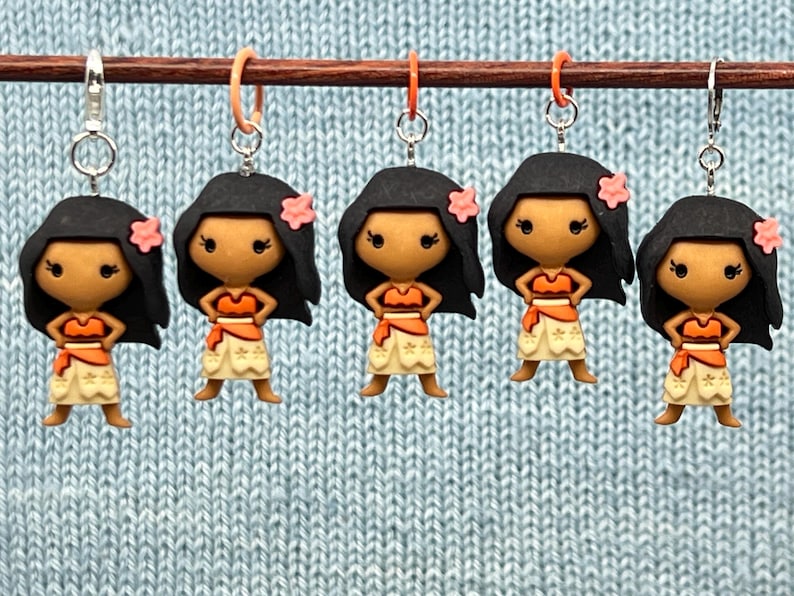 Puede incluir: Cinco marcadores de punto de ganchillo coloridos con un personaje de dibujos animados con cabello casta&ntilde;o, un corpi&ntilde;o marr&oacute;n, una falda amarilla y una flor rosa. Los marcadores de punto est&aacute;n colgados de una varilla de madera.