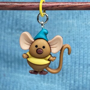 May include: A whimsical, cartoon-style mouse charm, featuring a brown body, large tan ears, and a blue hat. The mouse wears a yellow shirt and has a long, curved tail. It is attached to a silver-colored ring and a yellow marker.
