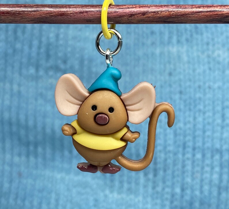 May include: A whimsical, cartoon-style mouse charm, featuring a brown body, large tan ears, and a blue hat. The mouse wears a yellow shirt and has a long, curved tail. It is attached to a silver-colored ring and a yellow marker.