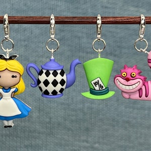 May include: Four Alice in Wonderland themed charm pendants. The charms are a purple teapot with black and white diamond pattern, a green top hat with a playing card that says "10/6", a pink Cheshire Cat with a wide grin, and a blonde Alice in a blue dress.