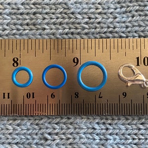 May include: An assortment of blue rubber O-rings of varying sizes and a silver-colored lobster clasp are displayed next to a ruler. The O-rings range from approximately 0.25 inches to 0.75 inches in diameter. The items are on a gray knitted surface.