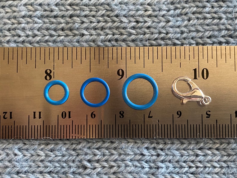 May include: An assortment of blue rubber O-rings of varying sizes and a silver-colored lobster clasp are displayed next to a ruler. The O-rings range from approximately 0.25 inches to 0.75 inches in diameter. The items are on a gray knitted surface.