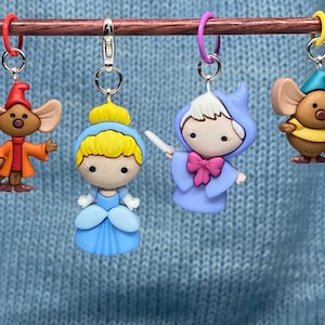 May include: Four colorful stitch markers featuring characters from a fairy tale. The markers include a mouse in an orange coat, Cinderella in a blue dress, a fairy godmother in a blue gown, and a mouse in a yellow shirt. Each marker has a metal clasp.