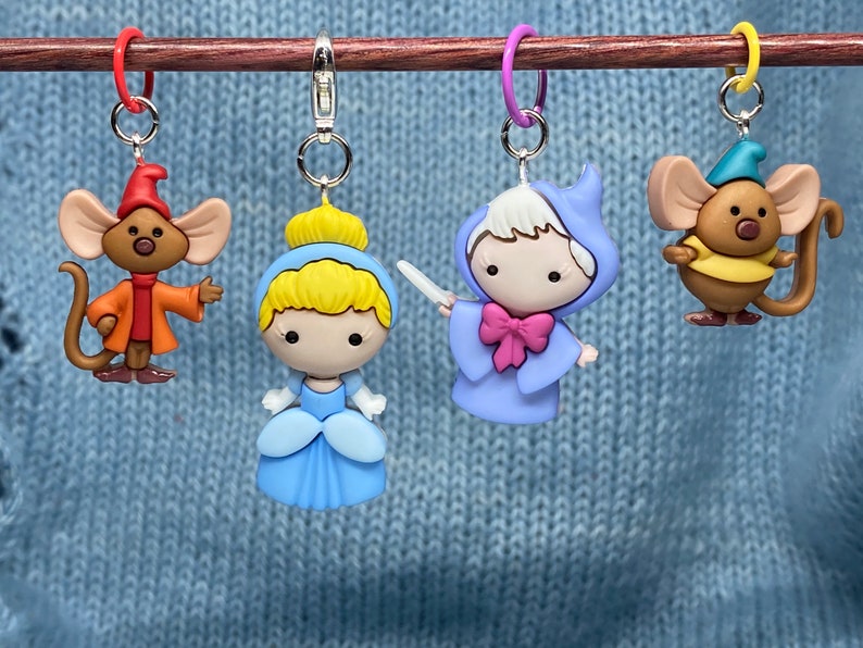 May include: Four colorful stitch markers featuring characters from a fairy tale. The markers include a mouse in an orange coat, Cinderella in a blue dress, a fairy godmother in a blue gown, and a mouse in a yellow shirt. Each marker has a metal clasp.