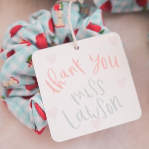 May include: A white card with the text "thank you miss Lawson" in pink and gray lettering with small pink hearts. The card is attached to a blue and white scrunchie with red apples printed on it.