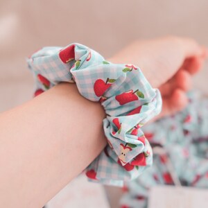 May include: A blue and white gingham hair scrunchie with red apple print.
