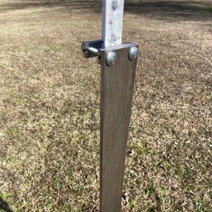May include: A metal sign holder attached to a white post with a clamp and bolts. The sign holder is a dark brown color and is mounted on a metal post. The background is a grassy field.