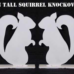 May include: Two white metal squirrel silhouettes, each standing on a white base. The squirrels are facing each other and are 8 inches tall. The text "8 IN TALL SQUIRREL KNOCKOVER" is printed in white at the top of the image.