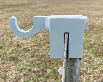 May include: A white metal hook attached to a metal post. The hook is shaped like a U and has a flat surface on the back. The post is painted white and has a rough texture.