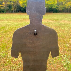May include: A metal silhouette target shaped like a person, standing on a metal pole in a grassy field. The target is a dark brown color and has a weathered appearance.
