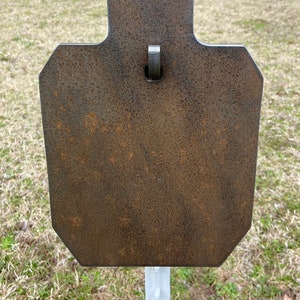 May include: A rusty metal shooting target with a rectangular shape and a small hook at the top. The target is mounted on a white metal post.