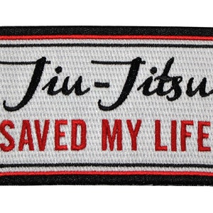 May include: A rectangular embroidered patch with a white background, a black border, and a thin red inner border. The text "Jiu-Jitsu" is stitched in black cursive script above the text "SAVED MY LIFE" which is stitched in bold red capital letters.