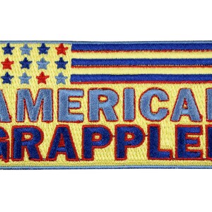 May include: A rectangular embroidered patch with a yellow background featuring a stylized flag design in the upper left corner with blue and red stars and stripes. The text AMERICAN GRAPPLER is embroidered in large blue letters with red borders.