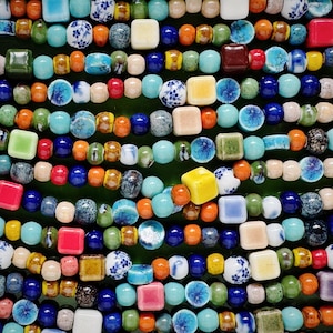 Könnte beinhalten: Nahaufnahme von bunten Perlen in einer grünen Schale. Die Perlen sind in Reihen angeordnet und umfassen verschiedene Formen und Farben, wie Blau, Orange, Gelb, Rot und Weiß. Einige Perlen haben florale Muster.