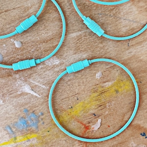 Puede incluir: Cuatro llaveros de cable turquesa con cierres plateados. Los llaveros están sobre una superficie de madera.