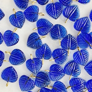 May include: A collection of cobalt blue glass leaf charms with gold-toned jump rings. The charms are all in the shape of a leaf with a slightly textured surface.